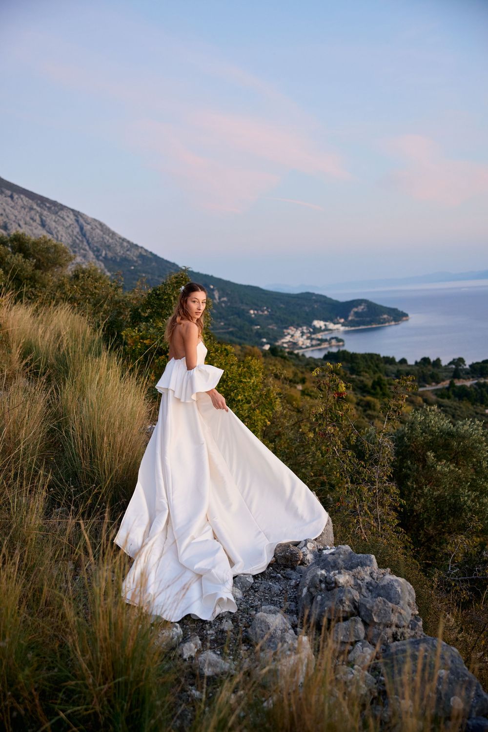 A bride in a flowing white wedding dress poses on rocky cliffs overlooking a scenic coastal mountain landscape at sunset.