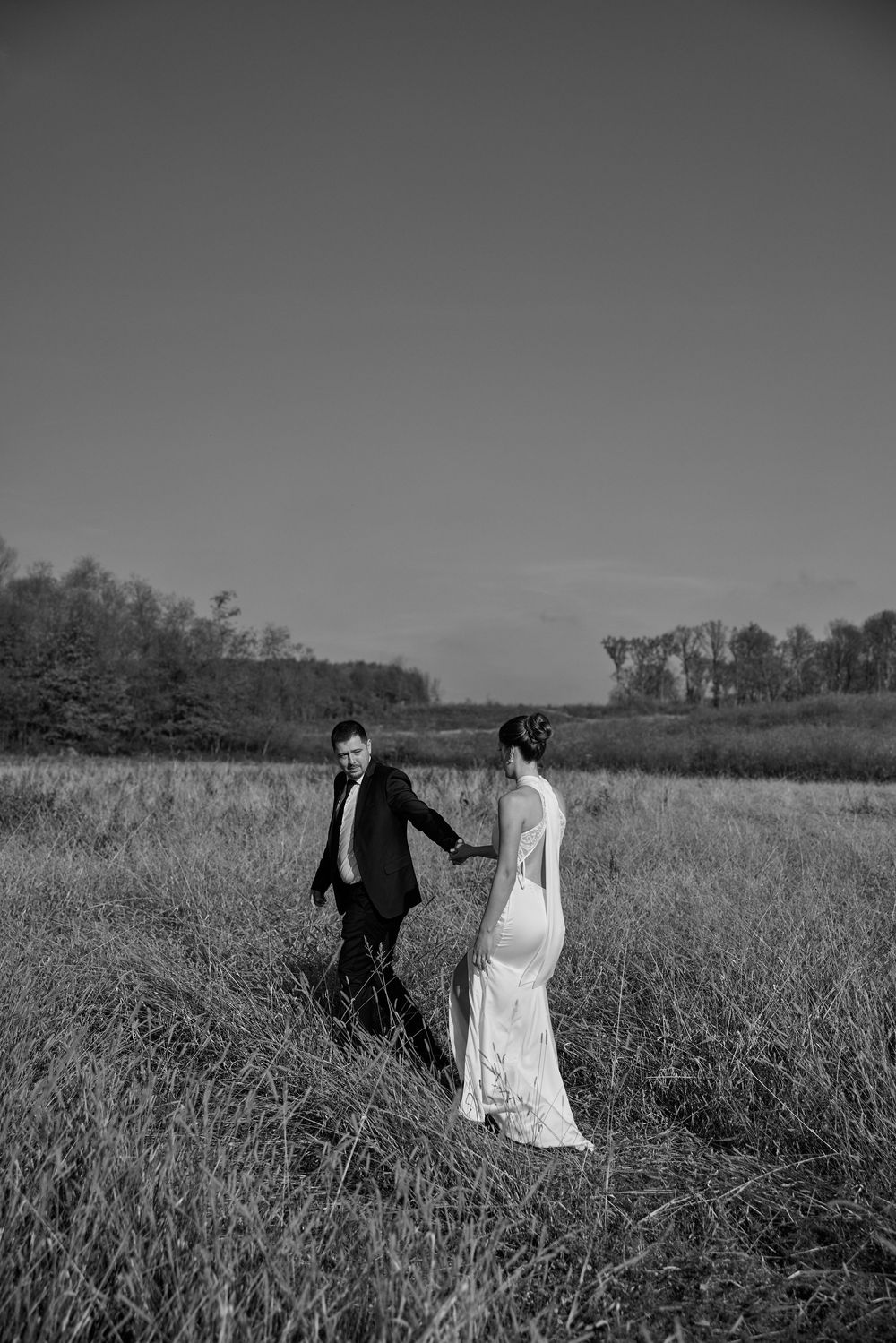 A couple in wedding attire walks together through a scenic field of tall grass in black and white.