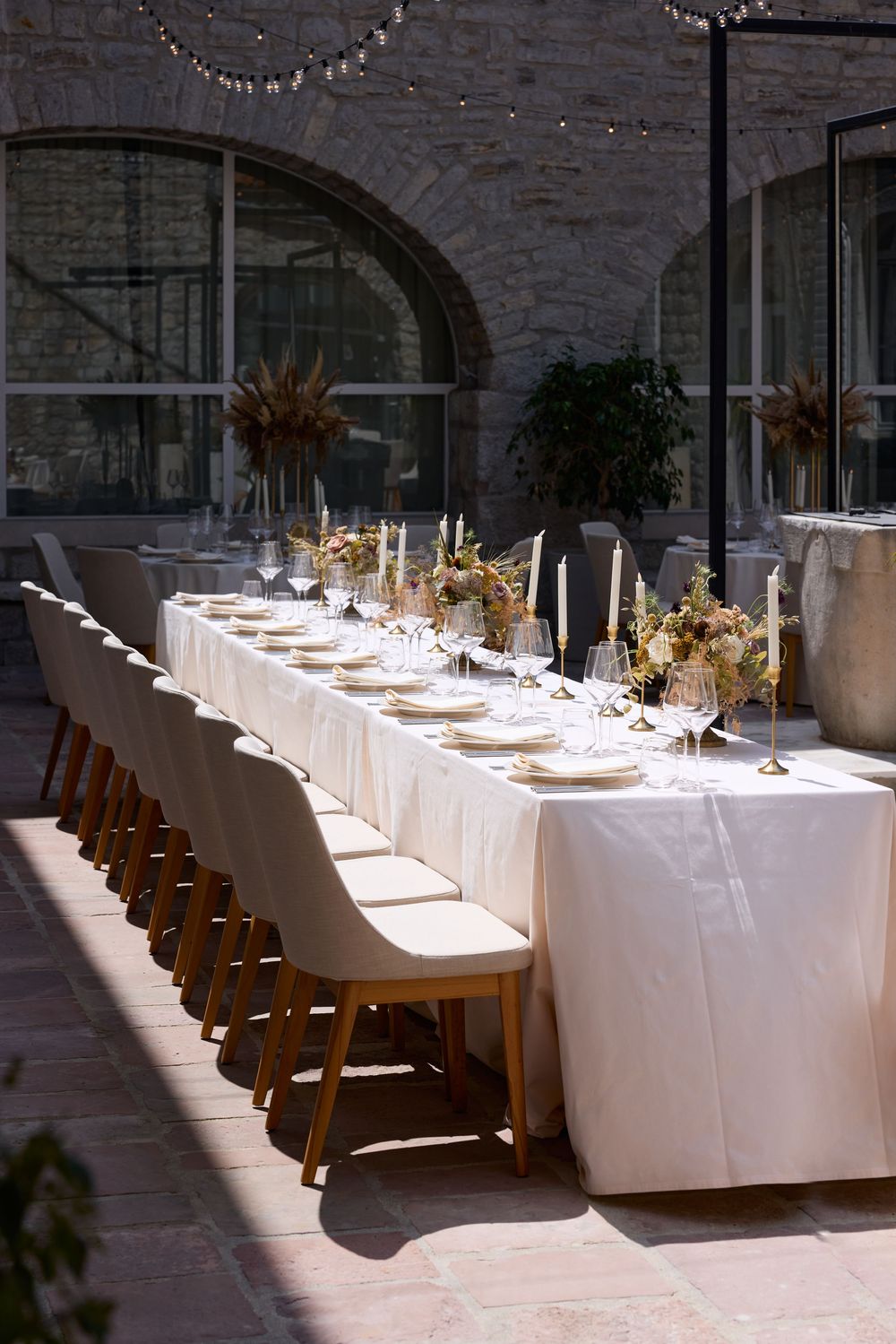 Long white banquet tables with wooden chairs set for an elegant indoor reception with natural lighting.