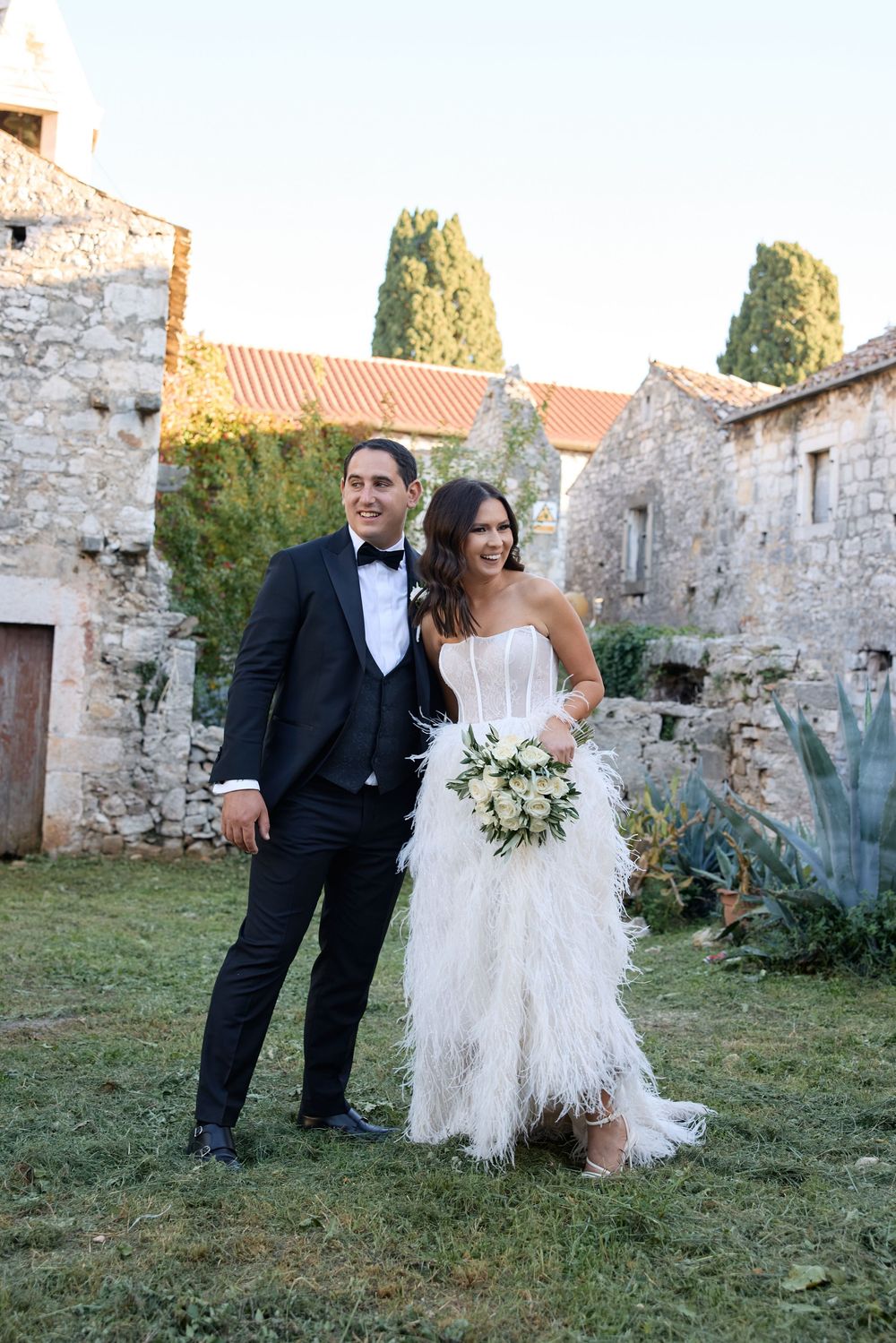 A wedding couple shares a romantic moment in front of rustic stone walls and gardens at sunset.