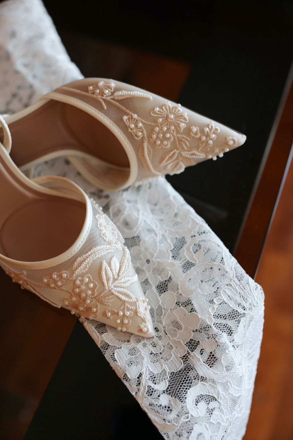 Elegant white bridal shoes with delicate floral beading and embroidery details rest on lace fabric.