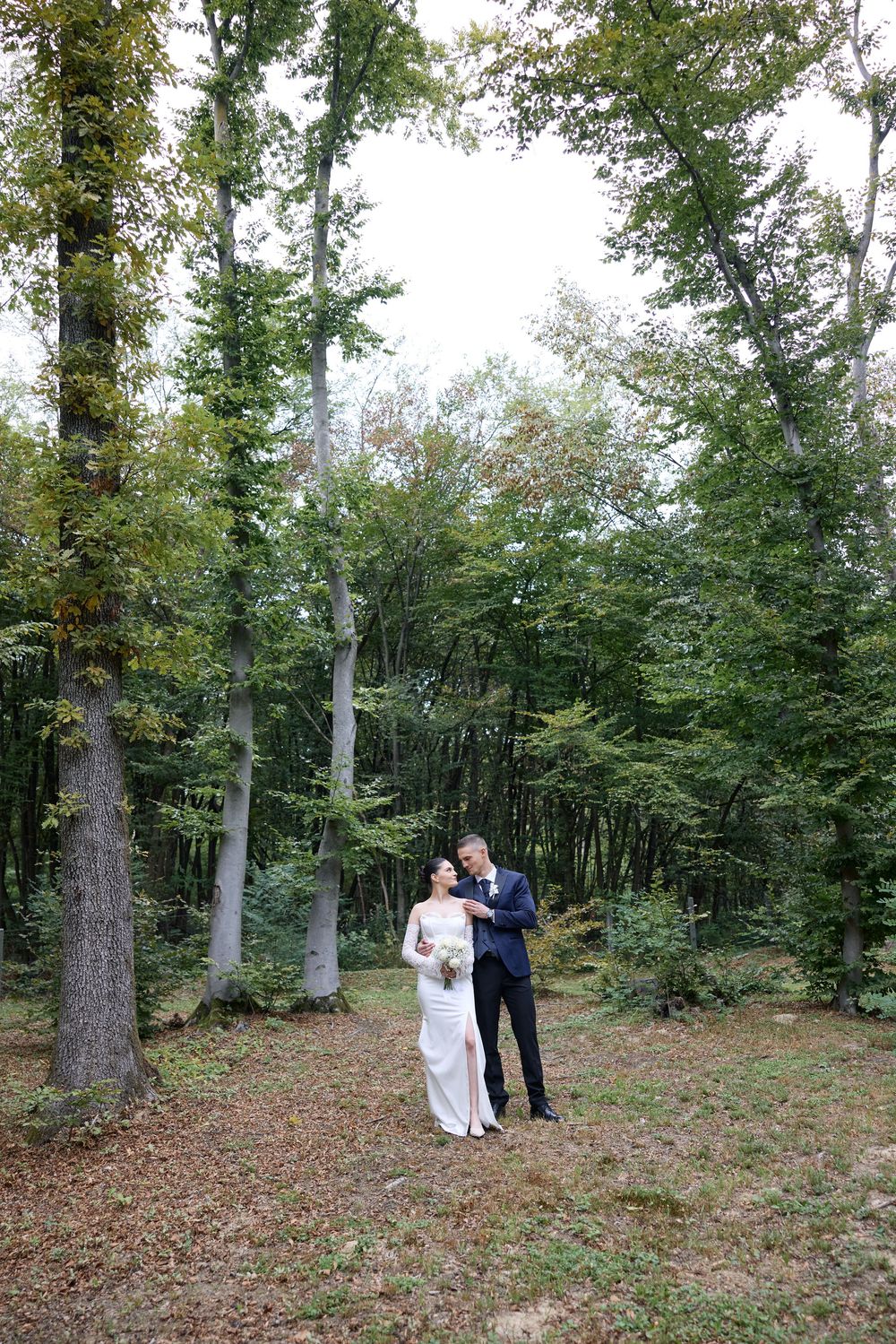 A romantic woodland photo session with a couple in wedding attire walking among tall trees and fallen leaves.