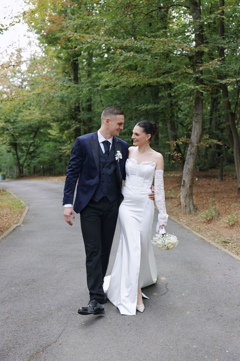 A newlywed couple walking together on a tree-lined path, with the bride in white and groom in black suit.
