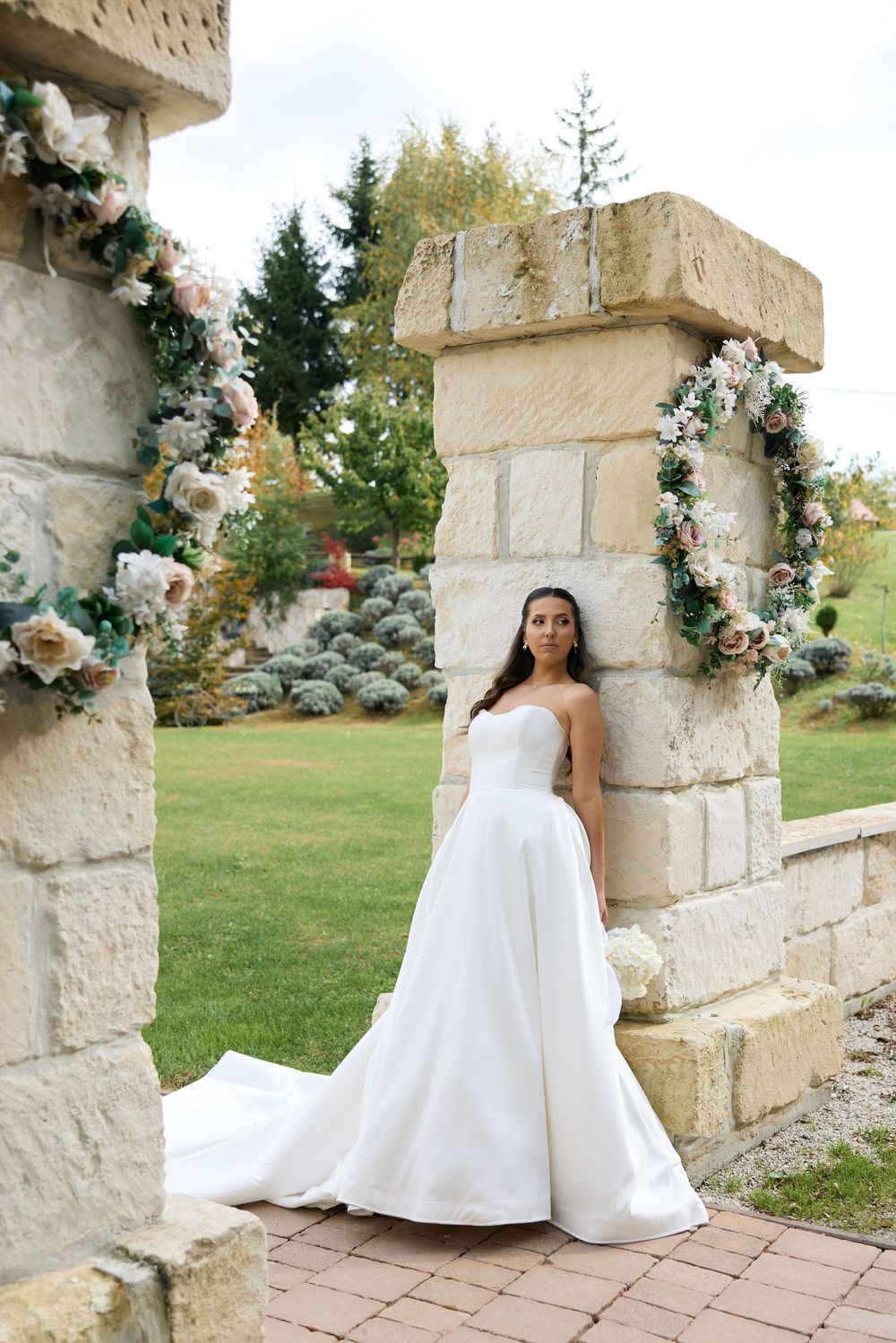 A white wedding dress is showcased against a rustic stone wall adorned with floral garlands in a garden setting.