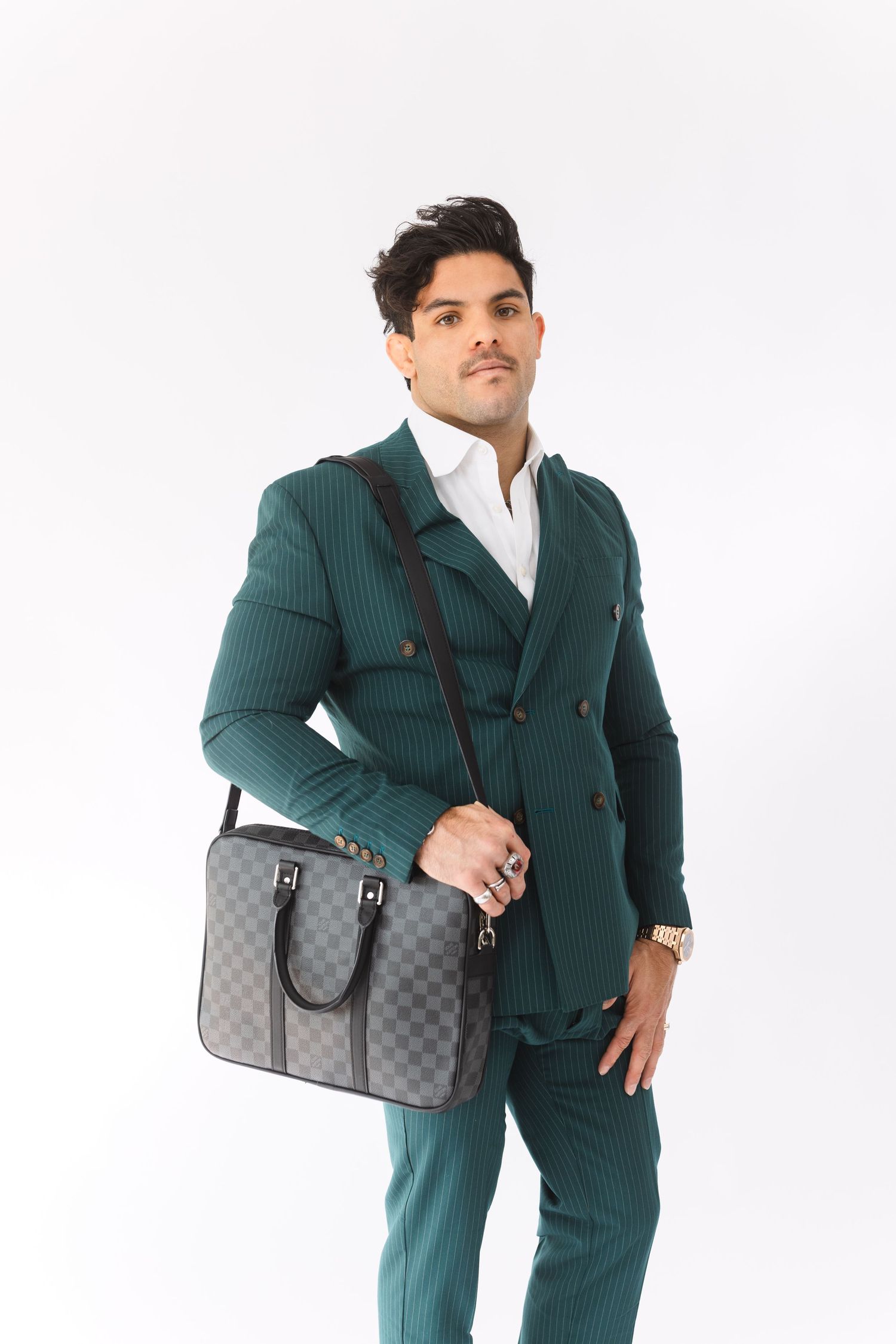 Professional model wearing a green pinstriped suit poses with a designer checkered grey messenger bag against white background.