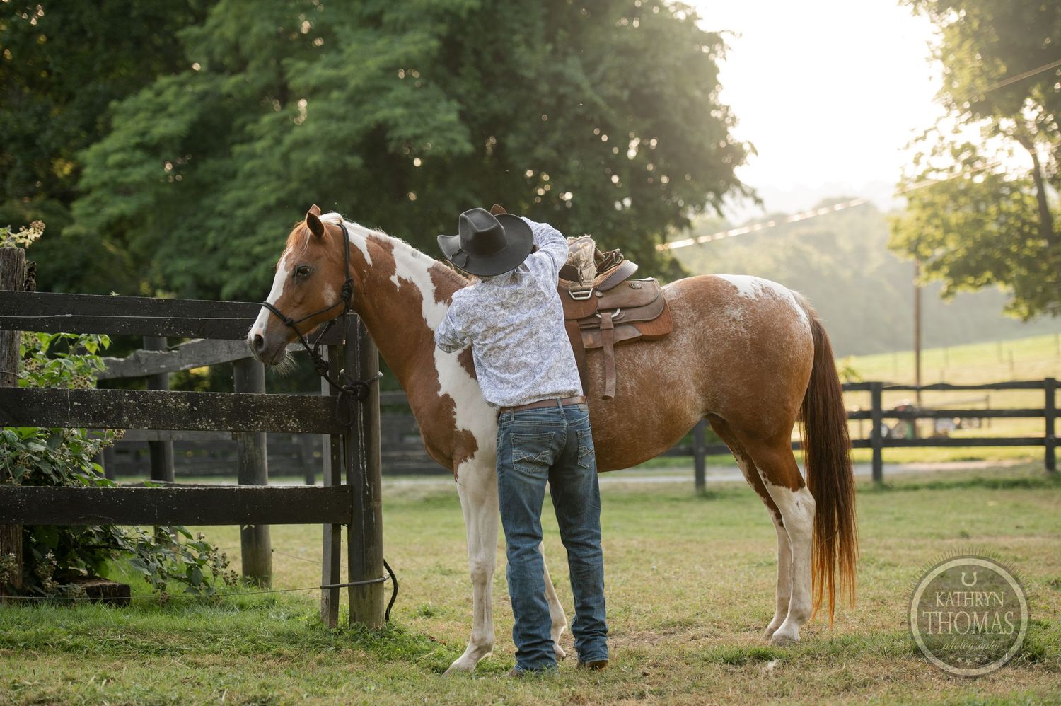 A person in western wear tends to a brown and white horse near a wooden fence at dusk.