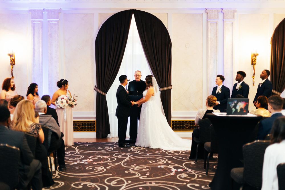 An indoor wedding ceremony takes place in an elegant hall with black curtains and ornate carpeting while guests observe.