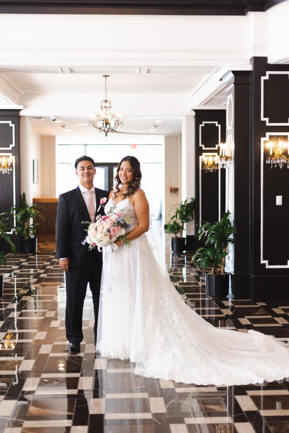 A couple stands elegantly in a luxurious hotel lobby with black columns, crystal chandeliers and patterned marble floors.