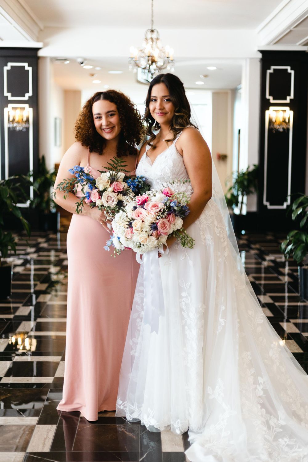 A bride and bridesmaid pose together in an elegant venue with black and white decor and chandeliers.