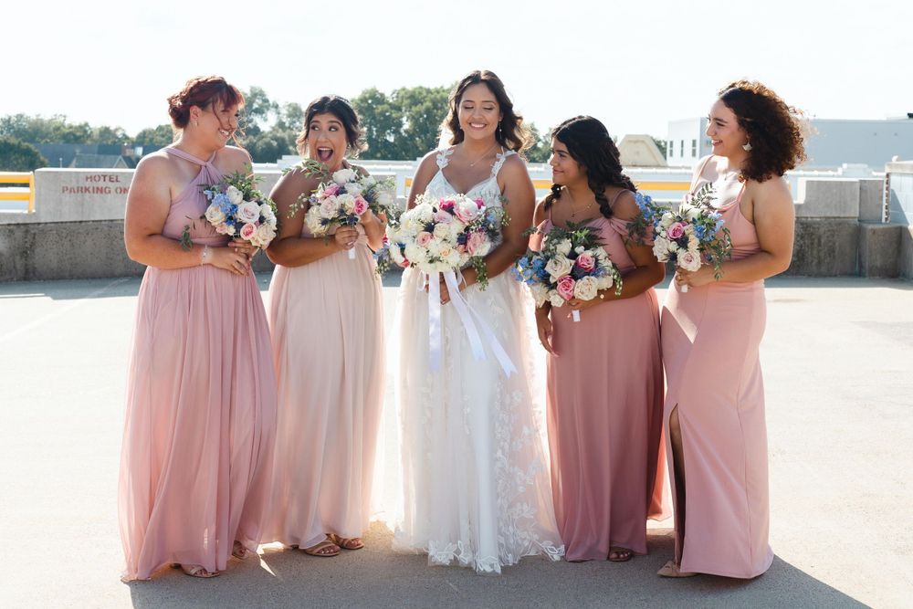 Bridesmaids in pink flowing dresses stand together with bouquets on a sunny outdoor patio.