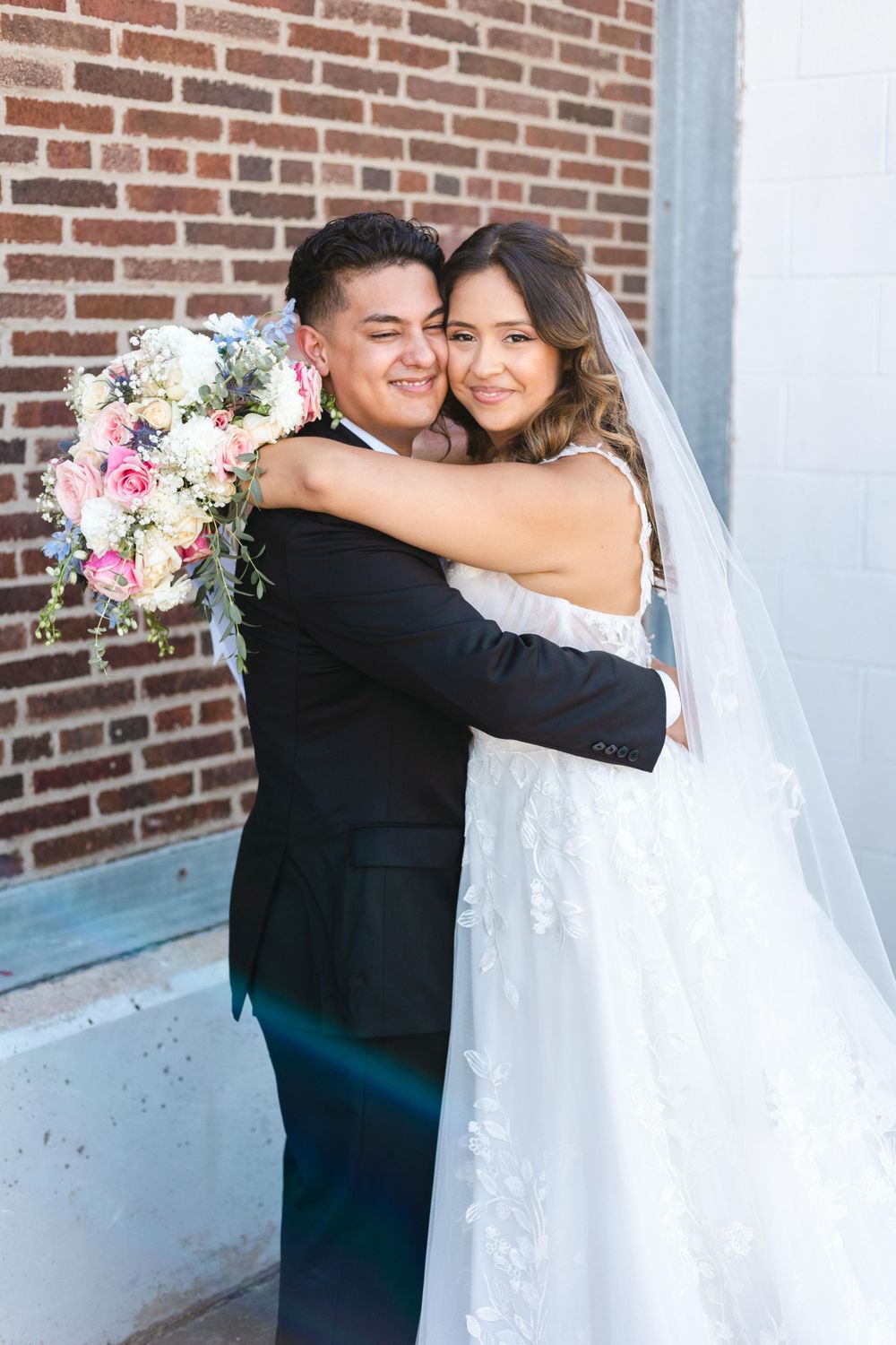 A newlywed couple shares an intimate moment against a brick wall backdrop.
