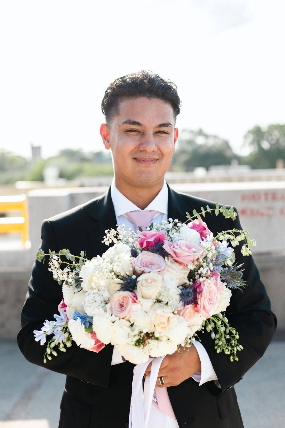 Wedding attendee in black tuxedo and pink bowtie holds beautiful pastel bouquet with roses and baby's breath outdoors.
