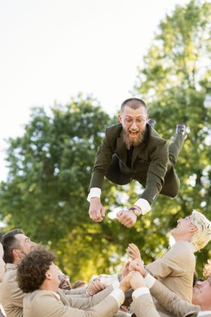 Wedding party members playfully lift and toss someone in a celebratory moment outdoors.