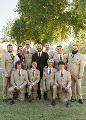 Groomsmen in tan suits pose together on grass for wedding photo.