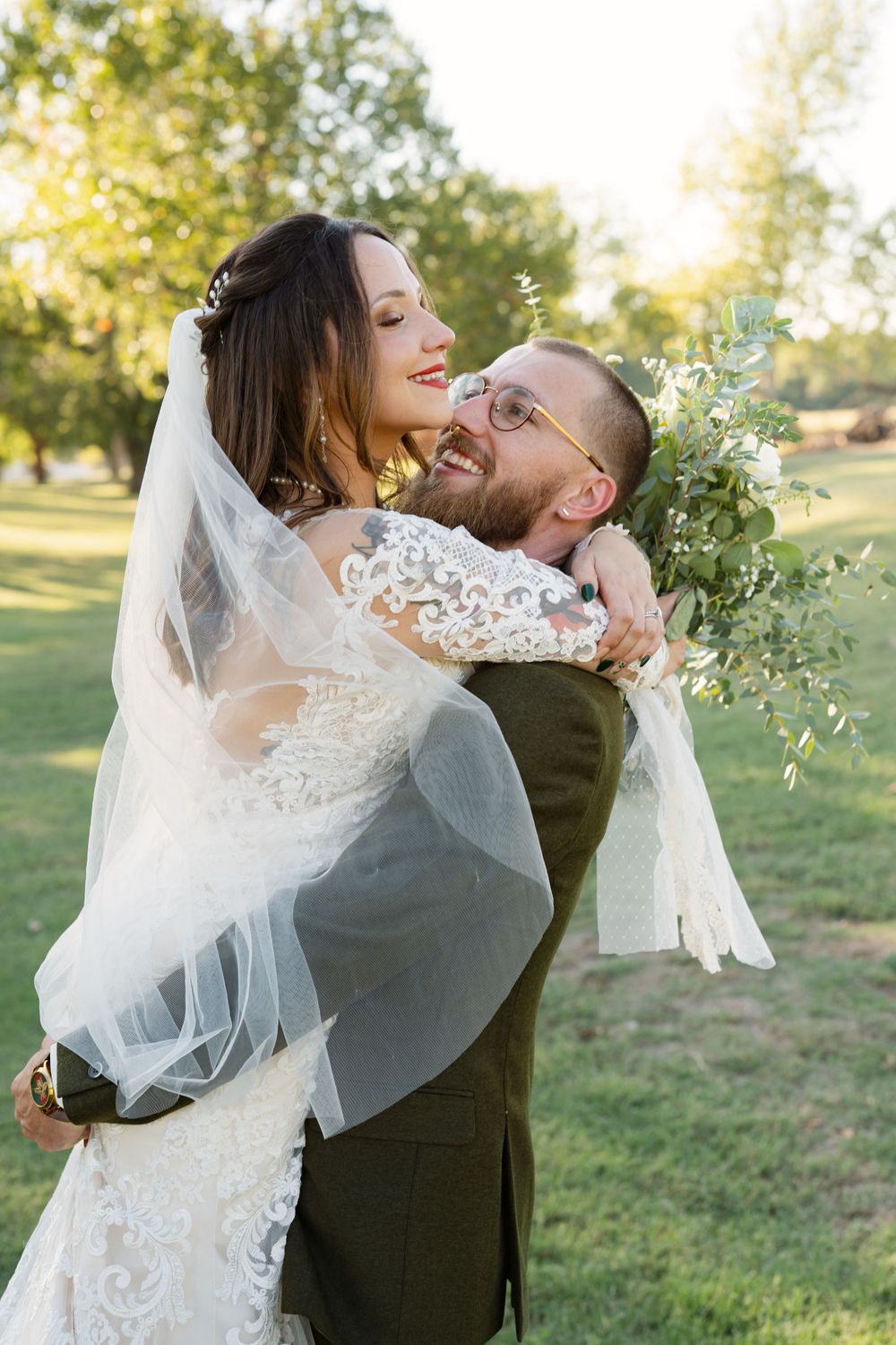 Joyful wedding couple embracing outdoors with flowing veil and greenery bouquet.