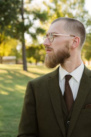 Professional portrait showing a well-dressed man in a dark suit with glasses and beard against an outdoor background.
