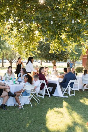 Wedding guests relax in white chairs under trees during an outdoor reception.