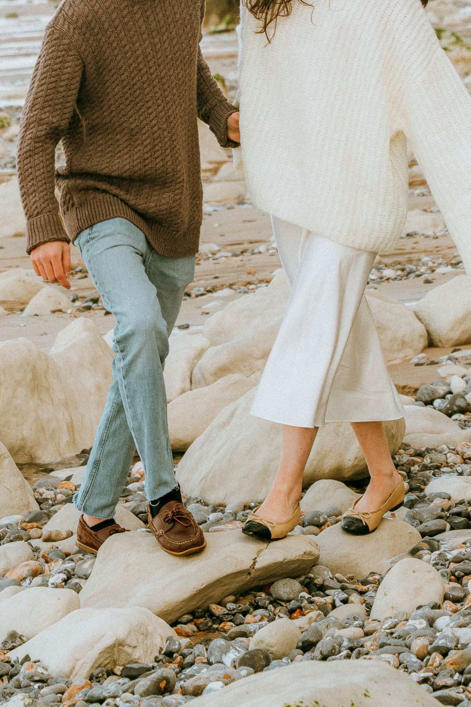 Two people wearing cozy knit sweaters and casual footwear stand together on rocky beach terrain.