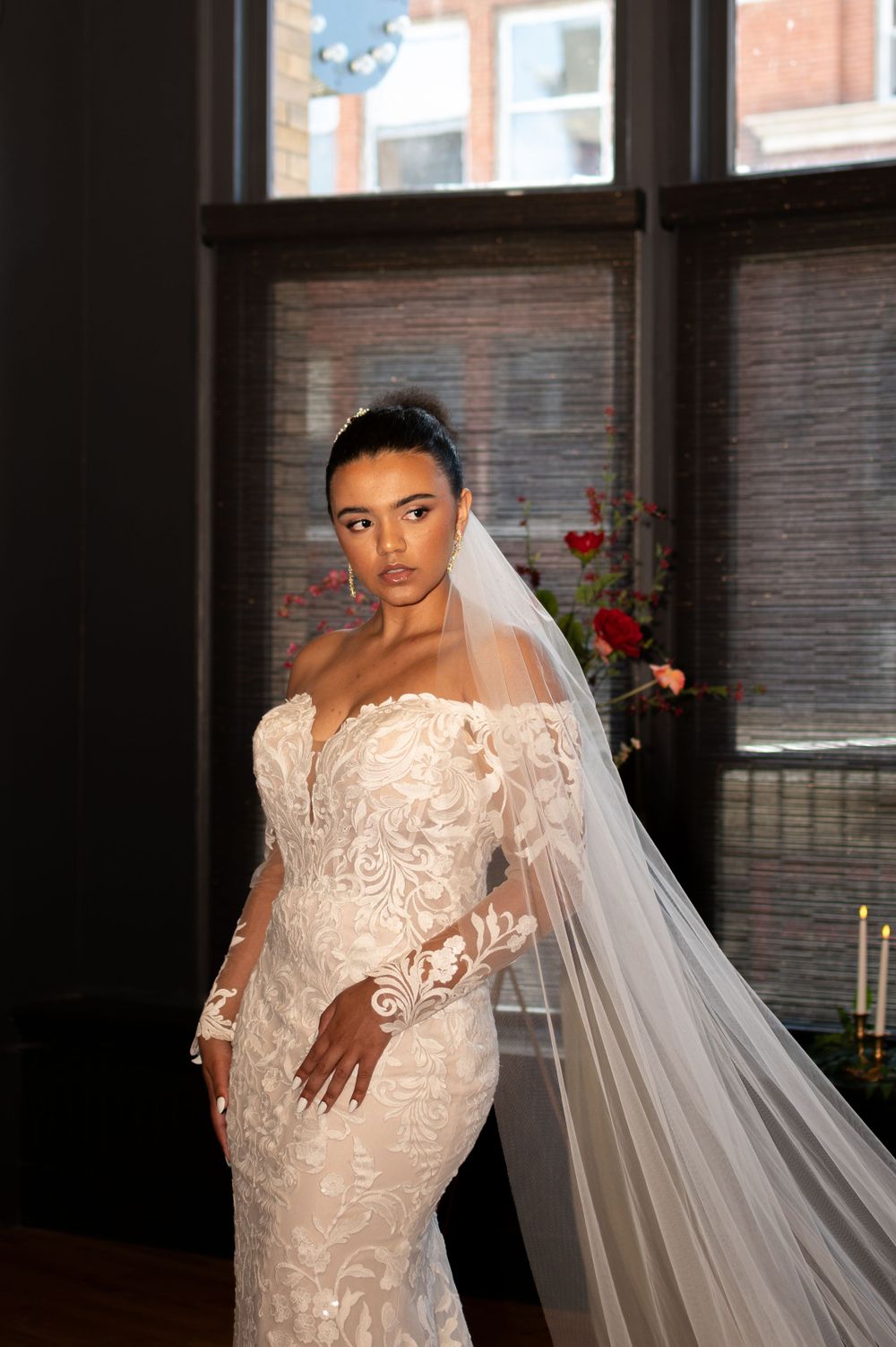 A bride wearing an elegant off-shoulder lace wedding gown with a long flowing veil poses in front of a window.