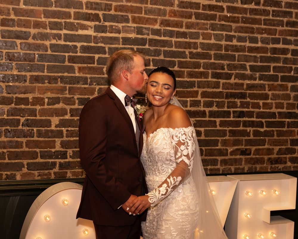 A couple shares an intimate moment in front of a brick wall with illuminated LOVE letters.