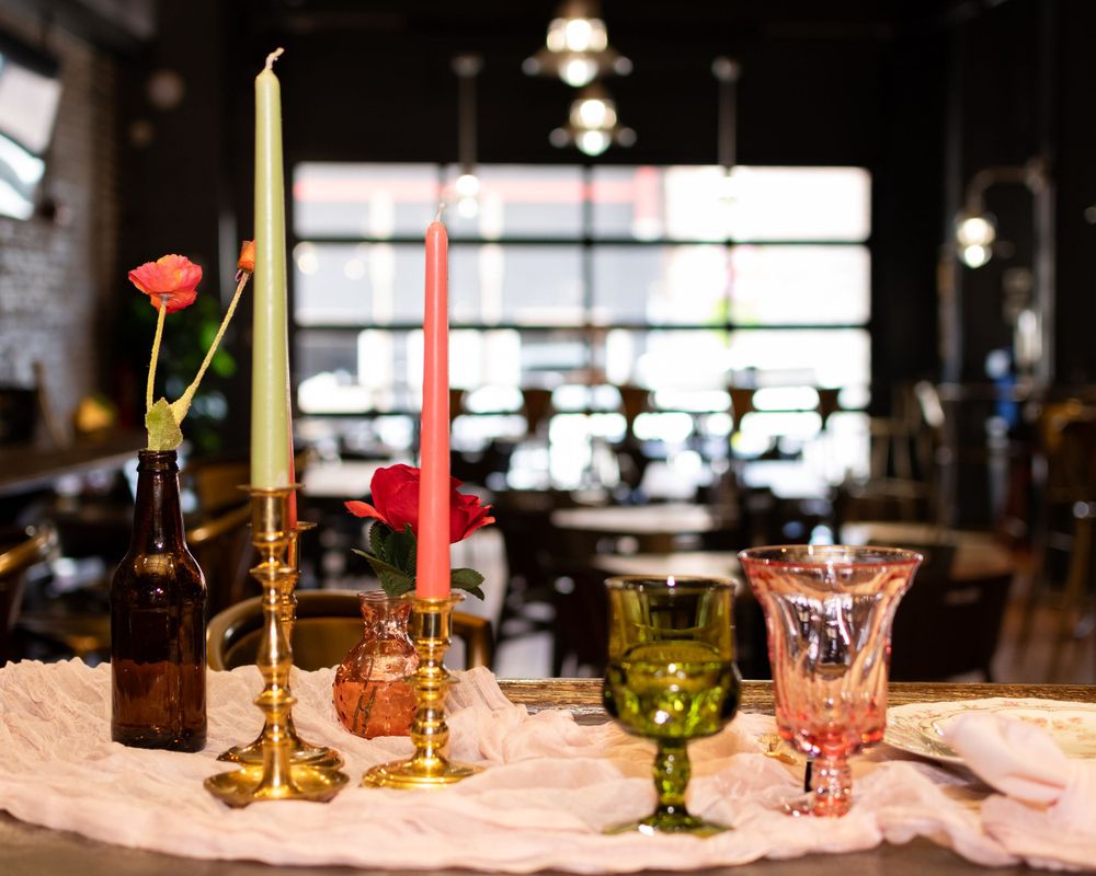 Colorful candles and vintage glassware arranged on a textured tablecloth in a dimly lit restaurant setting.
