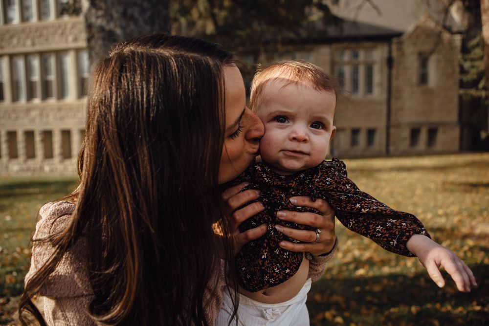 A parent giving a sweet kiss to their baby wearing a black lace shirt while standing in front of a brick building.