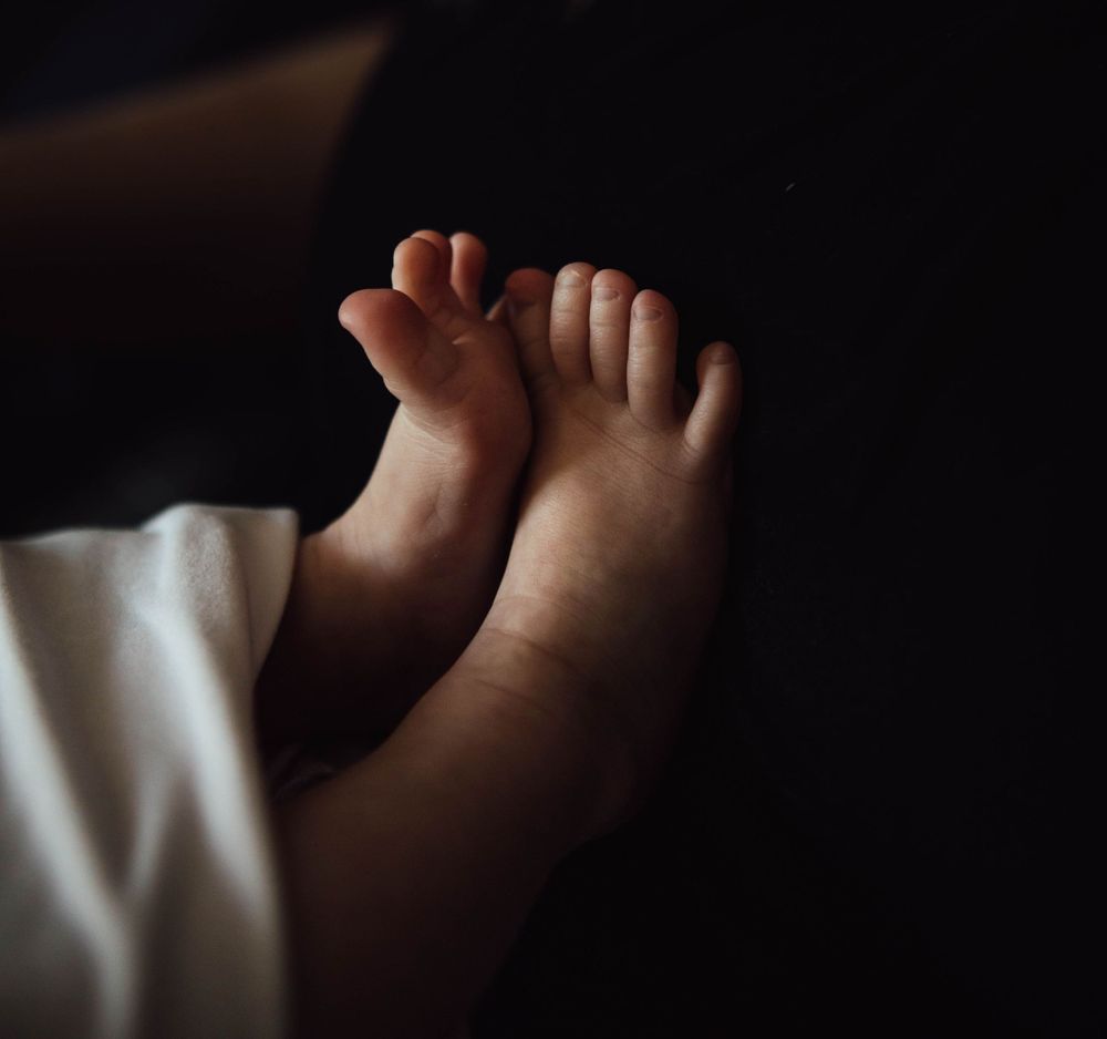Tiny newborn baby feet peek out from under soft white bedding in moody low-light photography.