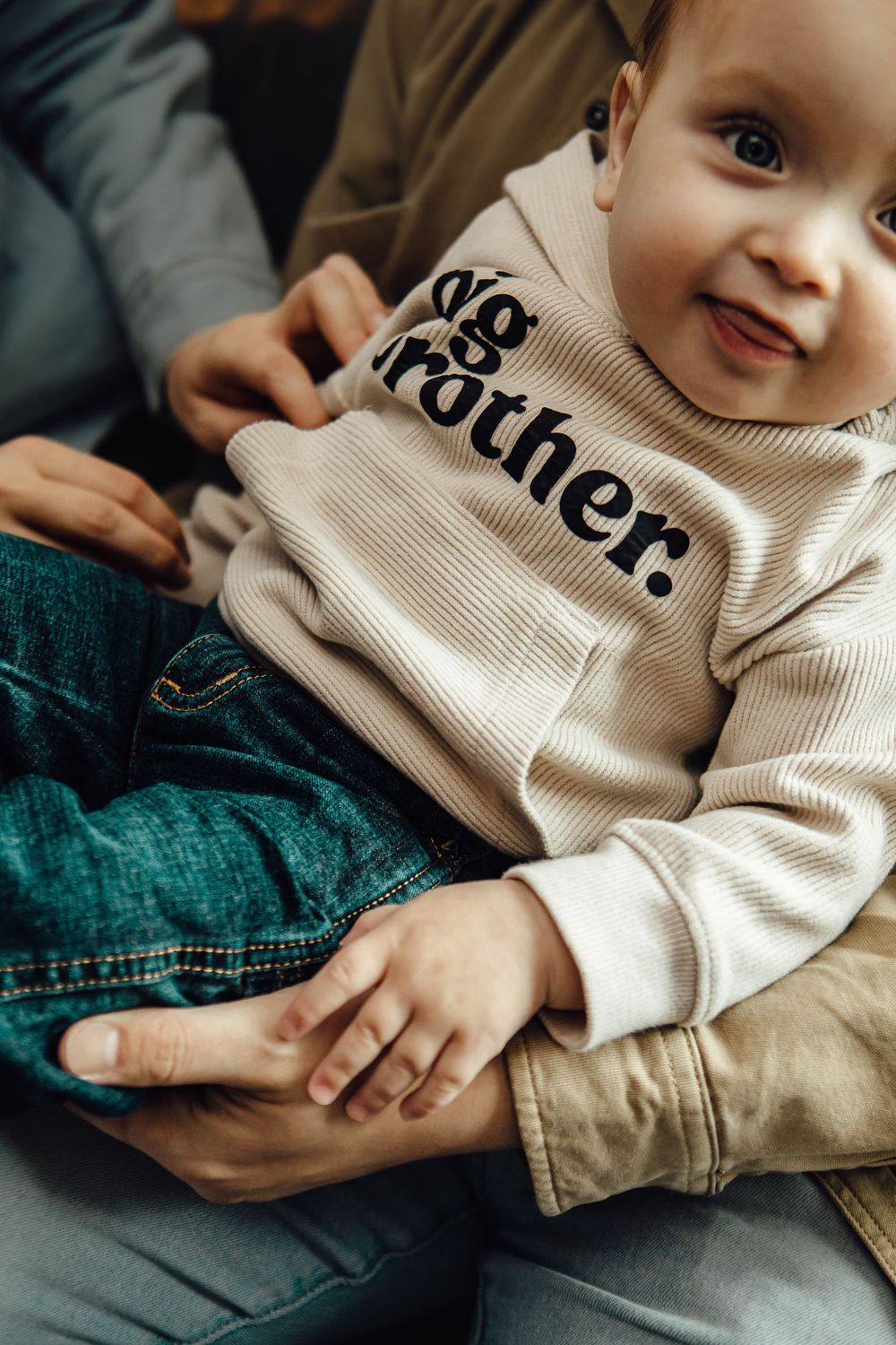 Baby wearing beige sweatshirt with 'Brother' text sitting with someone in denim jeans.