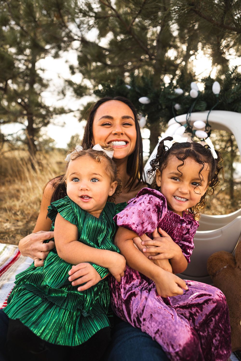 A joyful outdoor family portrait shows three people embracing and smiling together in a natural setting at golden hour.