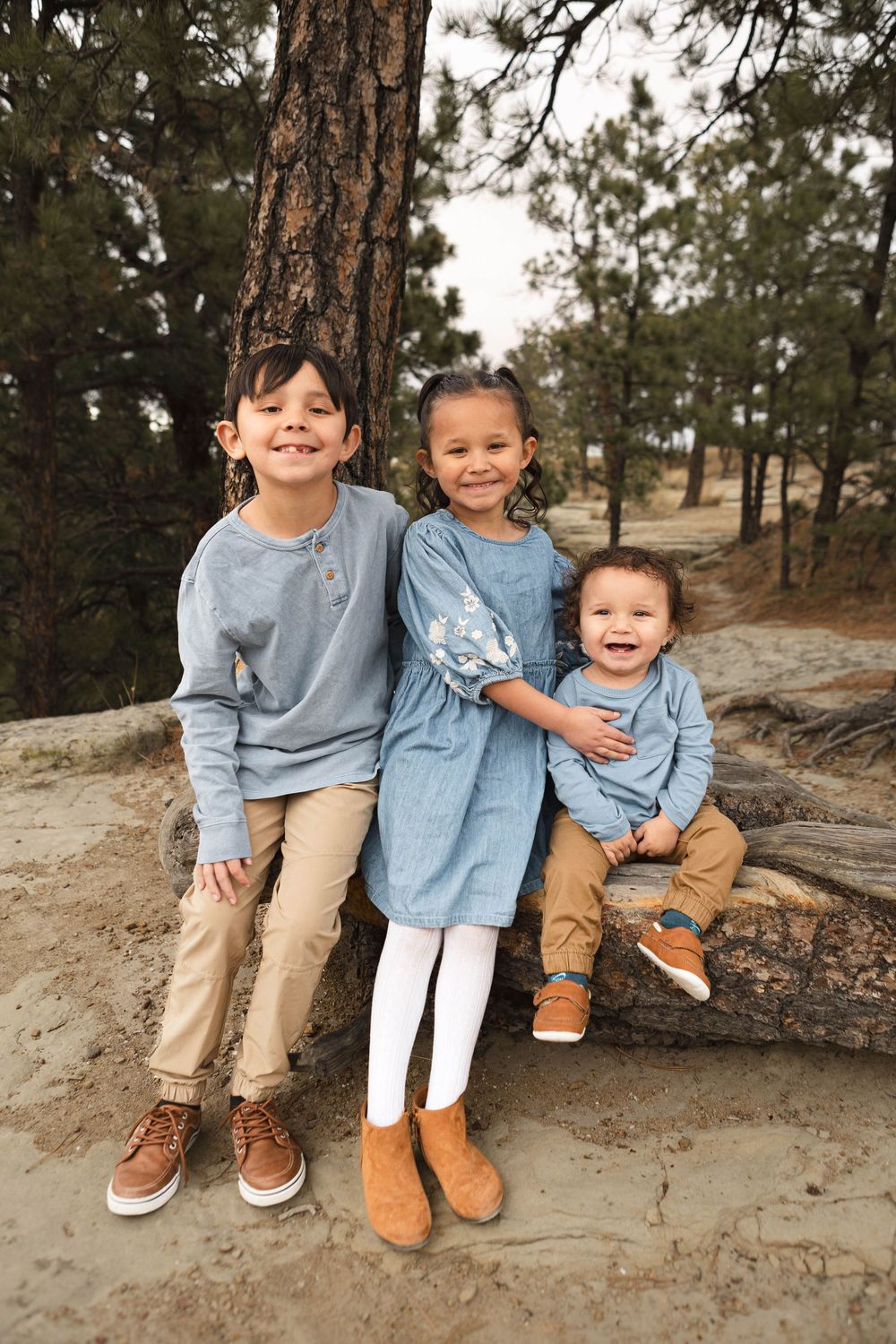 Three children in coordinating blue outfits sit together on a rocky ledge in a natural outdoor setting.