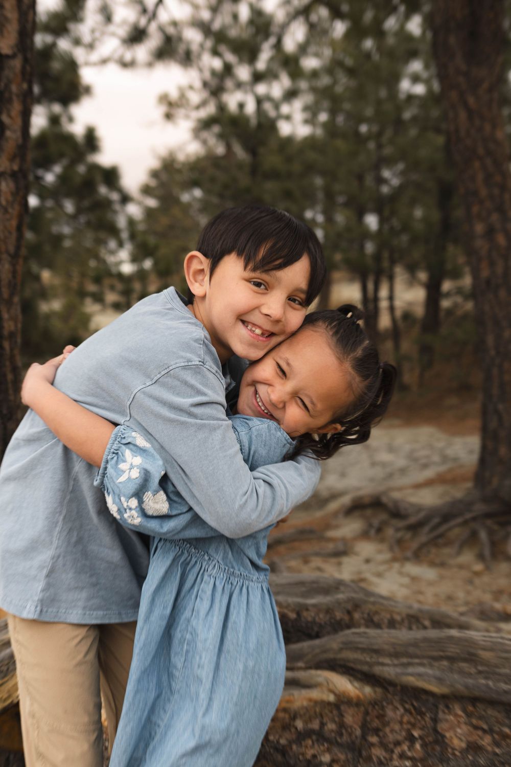 Two children in blue outfits share sweet hugs and laughs while playing outdoors among pine trees at sunset.