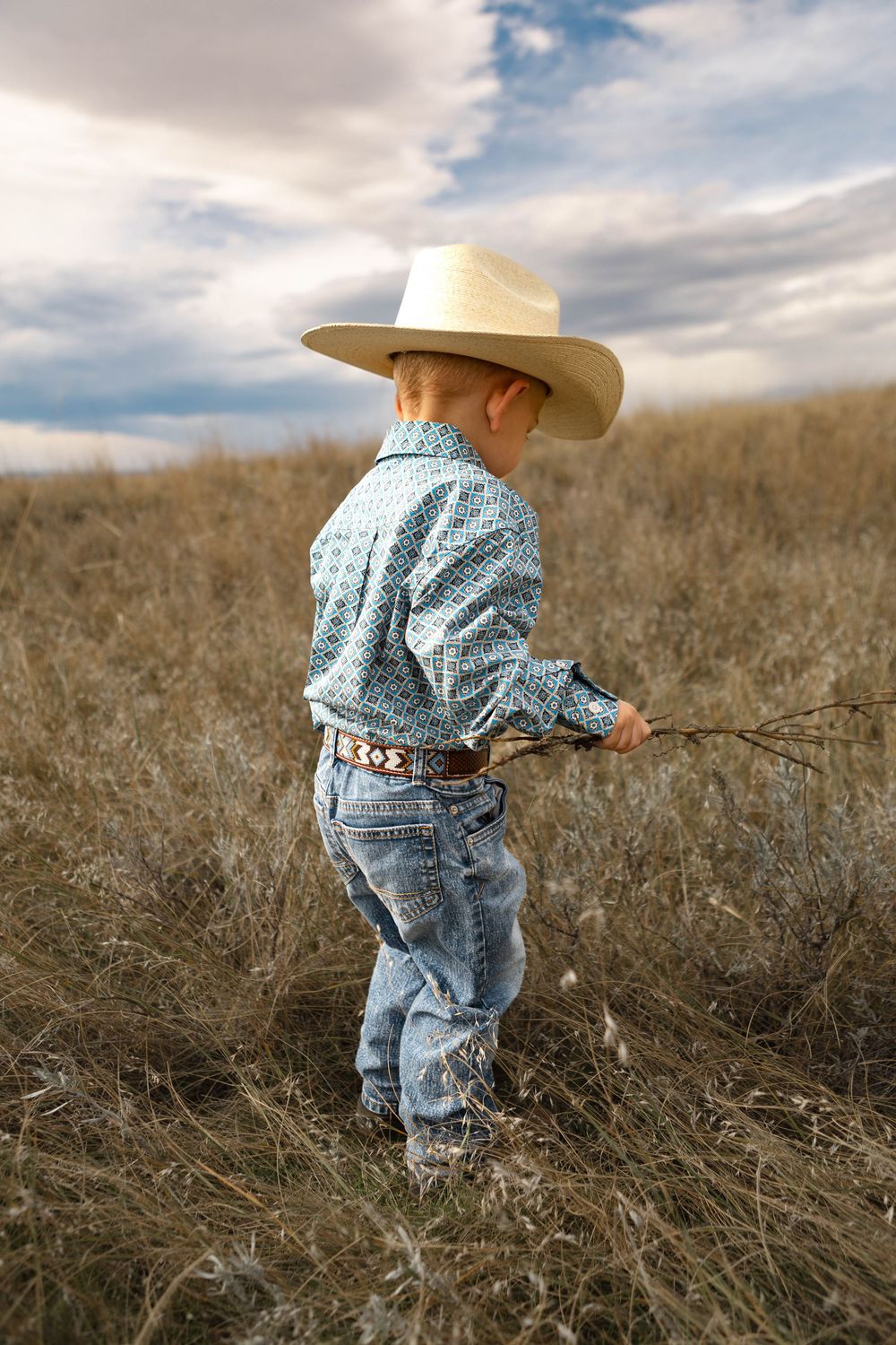 Young cowboy in blue shirt and jeans stands in a dry grass field under cloudy skies wearing a white western hat.