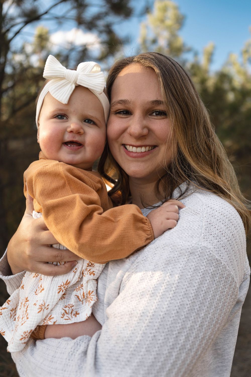 A heartwarming outdoor portrait shows a baby wearing a mustard sweater and white bow alongside someone in a white knit sweater.