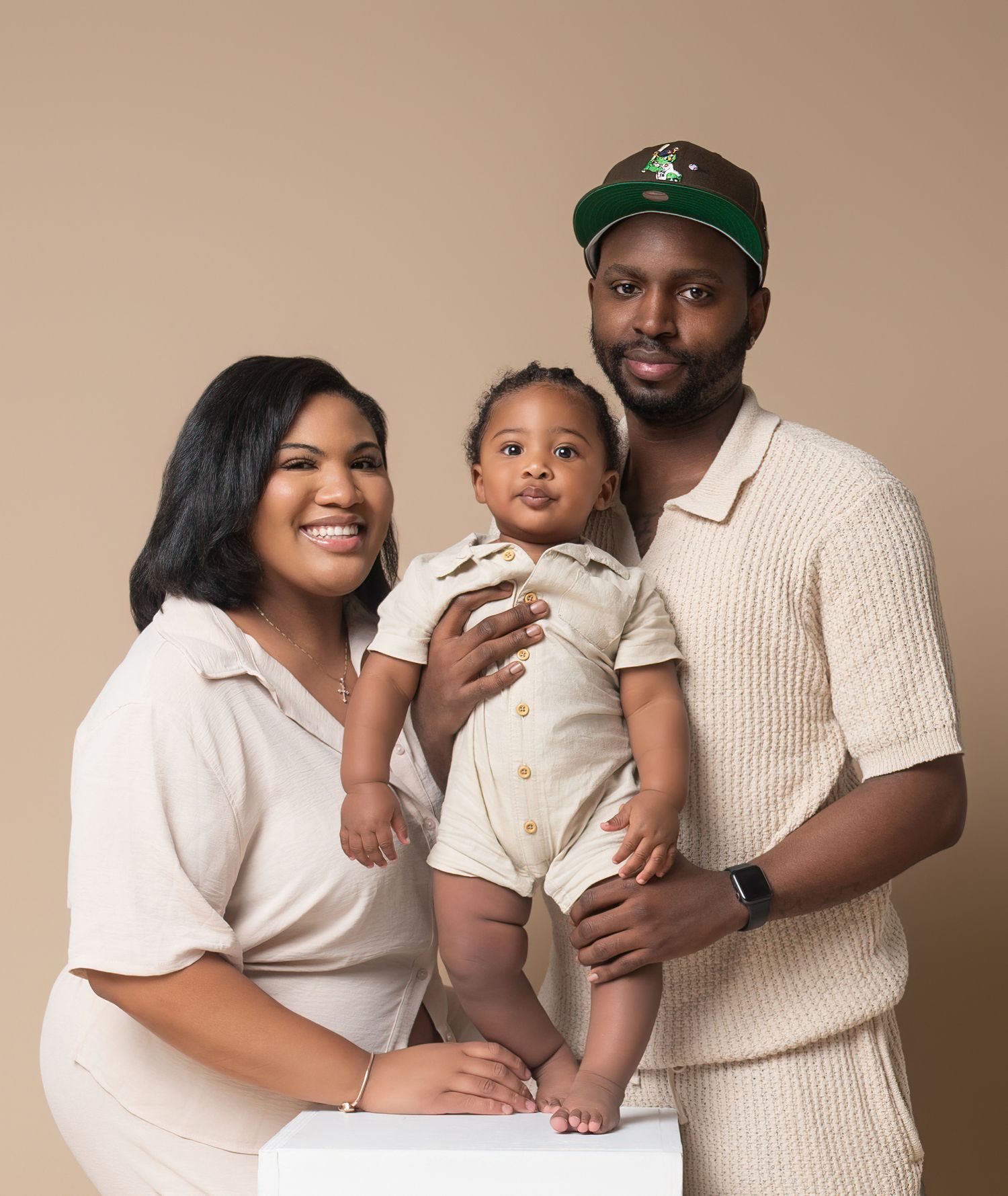 A loving family poses together in neutral cream-colored outfits against a warm brown backdrop for a studio portrait.