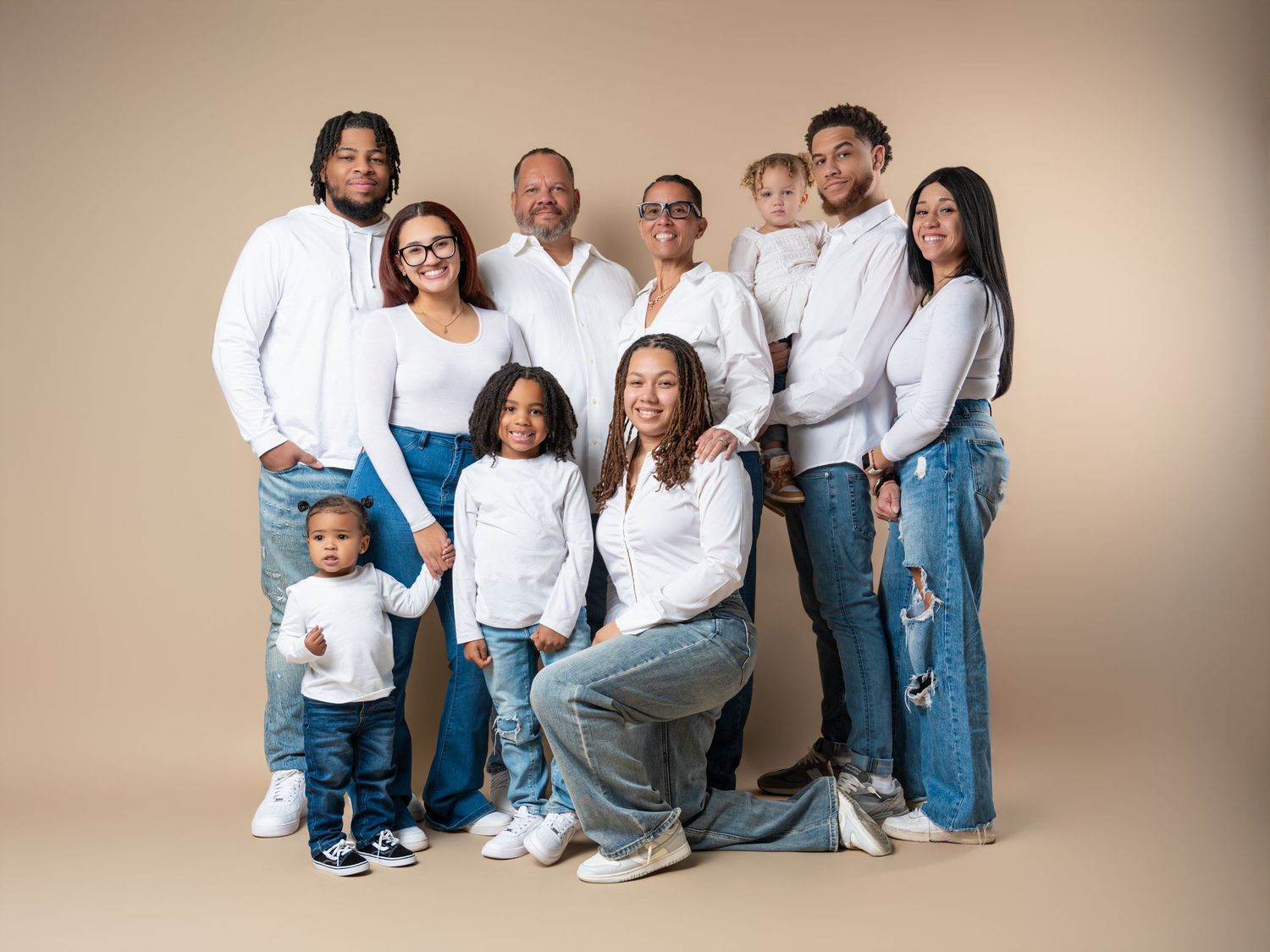Multi-generational family group wearing coordinated white tops and blue jeans poses together against beige backdrop.