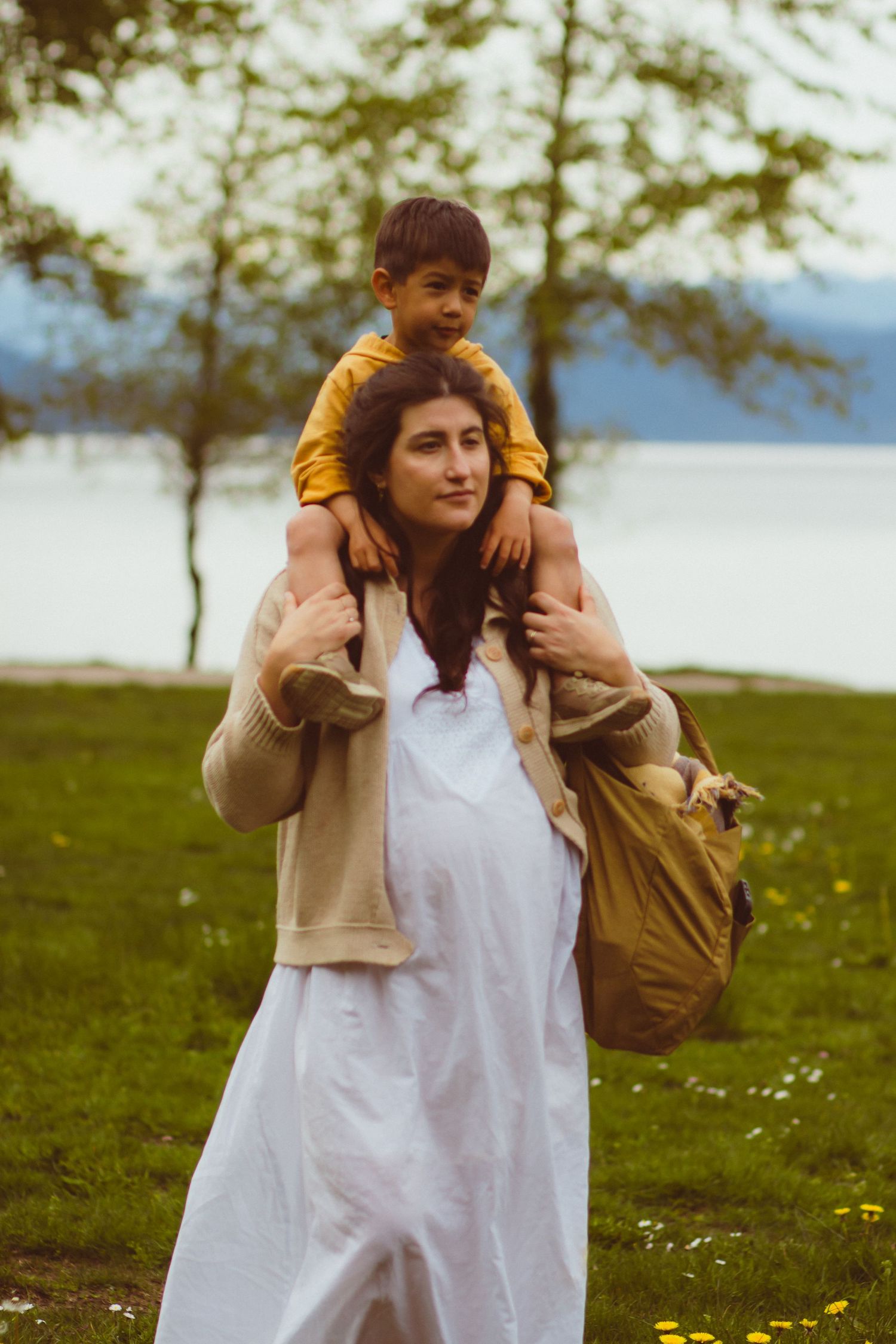 Mom in white dress showing her pregnant belly carrying her toddler boy on her shoulders after a family maternity photoshoot in Vancouver, BC