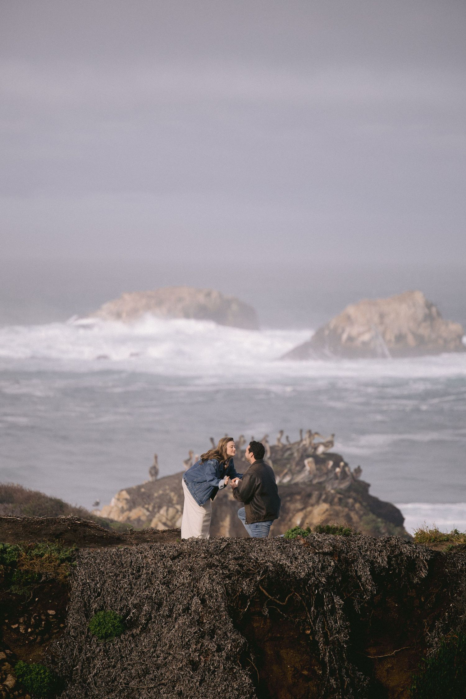 Couple sitting together on rocky cliff overlooking dramatic stormy ocean waves crashing against coastal rocks at sunset.