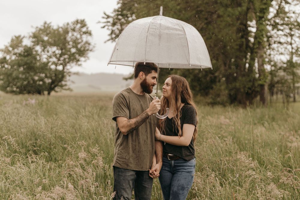 A romantic moment shared under a clear umbrella in a grassy field on an overcast day.