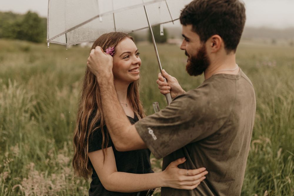 A joyful engaged couple shares laughter while holding a clear umbrella in a meadow.
