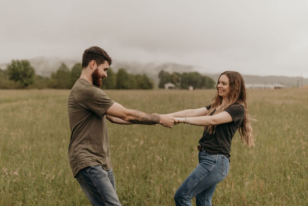 A playful engaged couple holds hands while spinning in a grassy field on an rainy day.