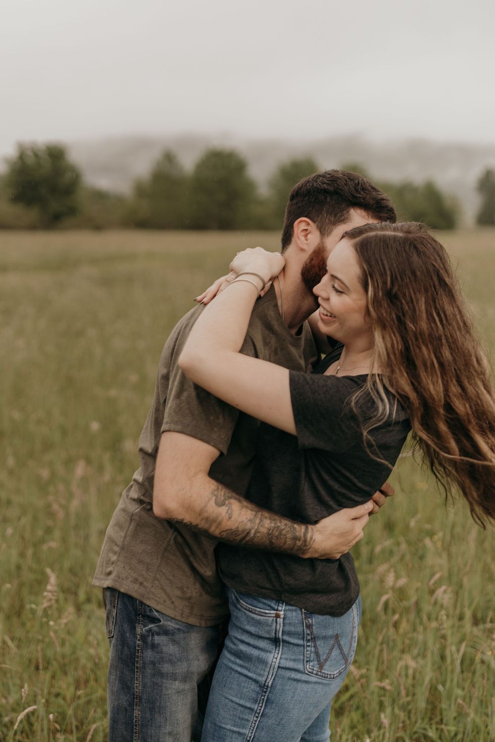 An intimate moment between a couple embracing in a rainy field with foggy hills in the background.