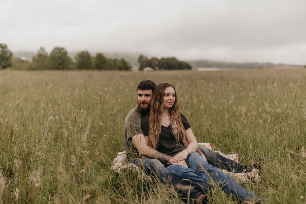 A couple relaxing together in tall grass during a rainy outdoor countryside egagement photoshoot.