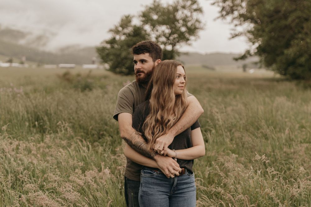A couple embraces in a serene meadow with rolling hills and trees in the background.