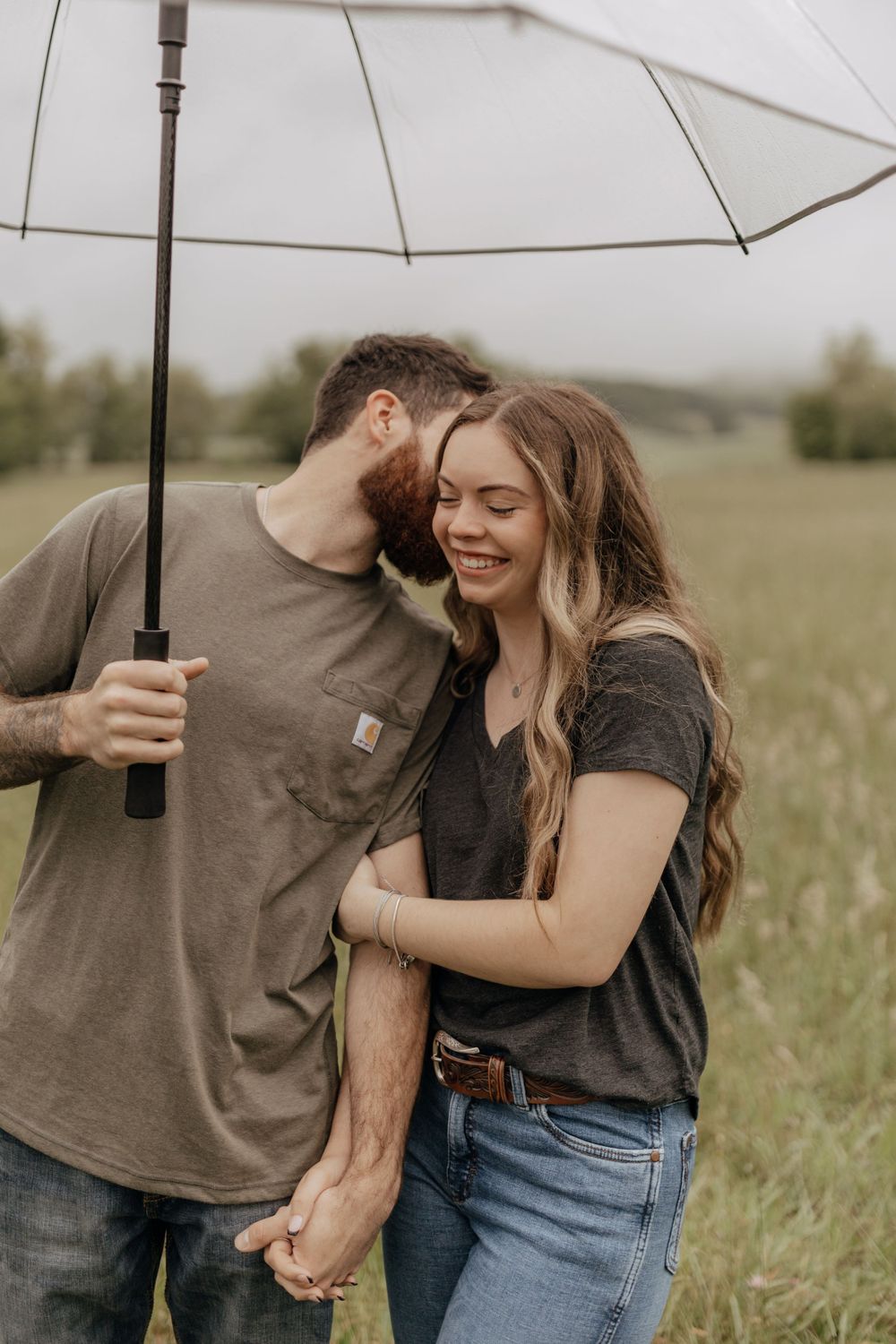 A couple shares an intimate moment under an umbrella in a grassy field.