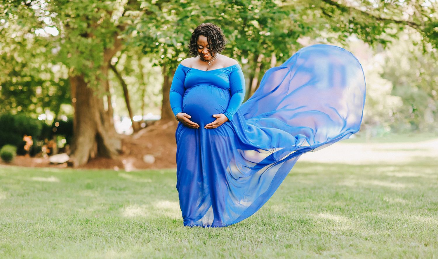 A maternity photo in a flowing blue dress with fabric billowing in the wind on a sunny day.