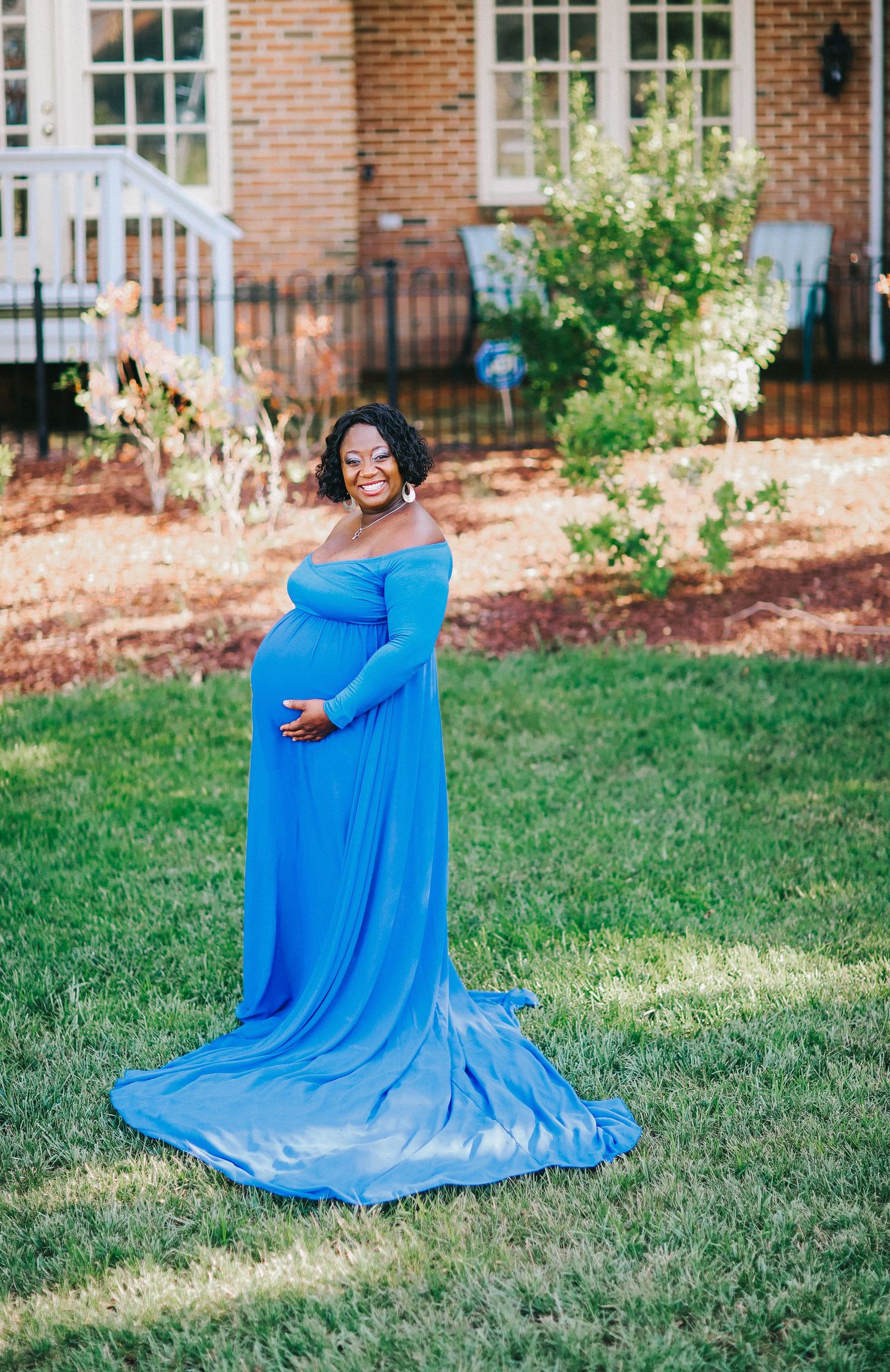 A maternity photo session outdoors with a flowing blue gown against brick house backdrop.