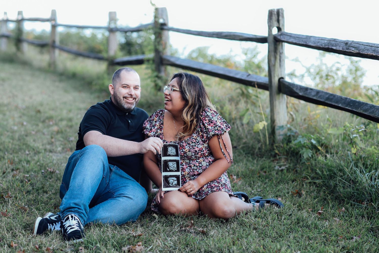 Outdoor maternity photo session with couple sitting by rustic fence holding ultrasound image.