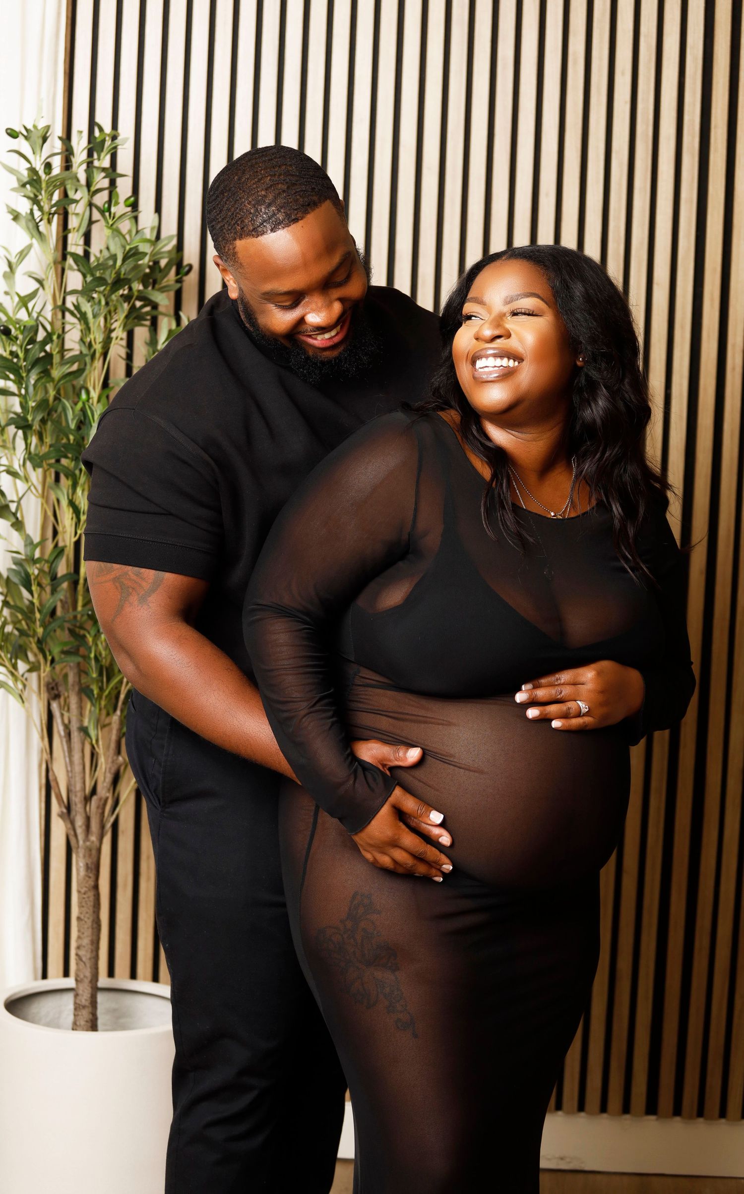 Pregnancy portrait featuring couple in black attire embracing in front of wood paneled wall.