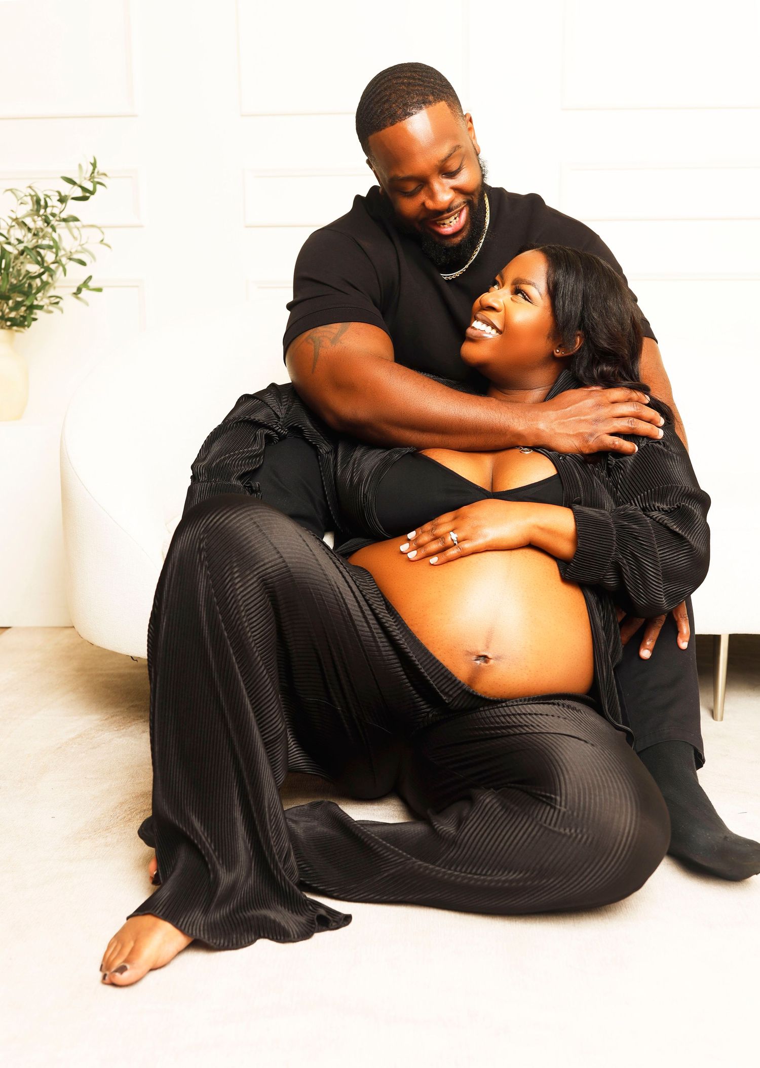 Intimate pregnancy photoshoot with couple dressed in black sharing tender moment on white floor.