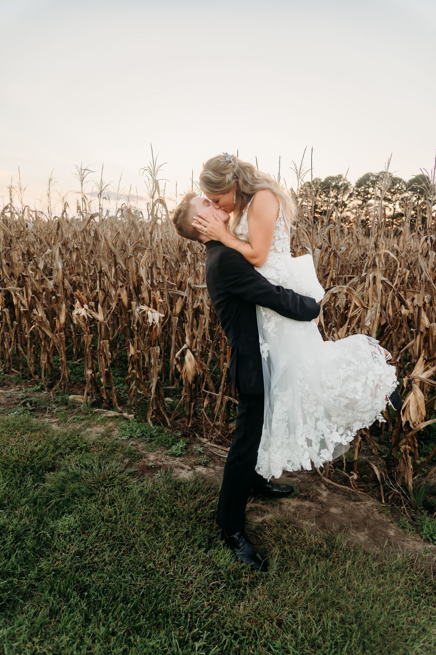 A romantic moment between newlyweds embracing by a cornfield at sunset as the groom lifts the bride in her white lace dress.